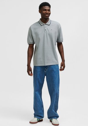 Ljusgrå pikétröja med krage, med en liten logotyp; kombinerad med lösa blå denimjeans och vita sneakers med gröna detaljer.