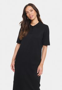 Melnā polo kleita, kas izgatavota no mīksta auduma, ar apkakli, īsām piedurknēm un pogu aizdari. Vienkāršs un elegants dizains.