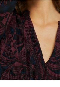 Bordeauxklänning med mörkblå blommönster. V-ringning och synlig veckad textur. Fokusera på tygdetaljer och färgkontrast.