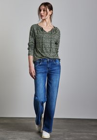Blusa lunga a maniche lunghe verde a fantasia con scollo a V, abbinata a jeans a gamba larga blu chiaro e sneakers bianche su un pavimento grigio.