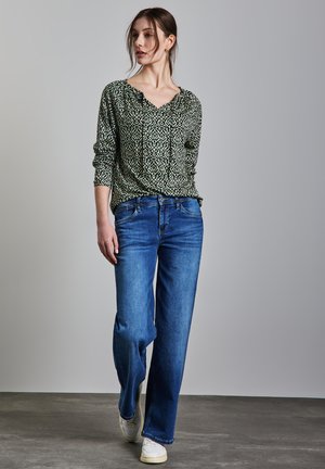Blouse longue à manches vert à motifs avec un décolleté en V, associée à un jean large bleu clair et des baskets blanches sur un sol gris.