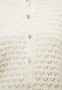 Πλεκτή κρεμ ζακέτα με διακριτικό μοτίβο σε σχήμα διαμαντιού, έξι ασημένια κουμπιά και υφή για επιπλέον λεπτομέρεια.