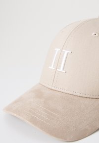 Beige Kappe aus strukturiertem Stoff. Mit weißer, gestickter „II“ auf der Vorderseite. Weiche, matte Oberfläche am Schirm und sichtbare Nähdetails.