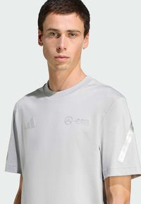 Grå atletisk T-shirt lavet af glat stof. Har et diskret logo og striber på ærmerne. Rund halsudskæring og løs pasform.
