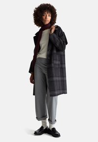 Tweedowy płaszcz we wzór kraty w ciemnoszarym kolorze z dużymi guzikami; w połączeniu z jasnym swetrem, szarymi spodniami i czarnymi mokasynami. Burgundowy szalik dodaje kontrastu.