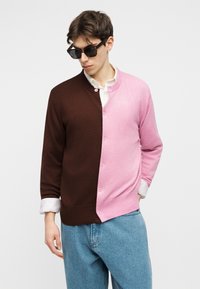 Vīrietis saulesbrillēs ar pusbrūnu, puspink kreklu ar pogām virs baltas krekla un zilām džinsu biksēm, ar roku kabatā.