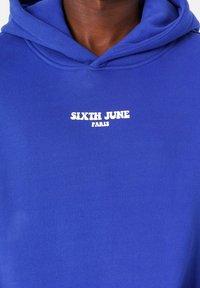 Sudadera azul hecha de tela suave con capucha ajustable. Presenta texto blanco "SIXTH JUNE PARIS" en el pecho. Diseño simple sin patrones.