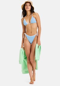 Light blue bikini with white trim; triangle top and tie-side bottom. Model holds a green sweater. Minimalist, smooth texture.
