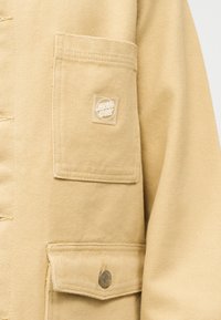 Béžová látková bunda s dvoma viditeľnými vreckami, jedno s výraznou nášivkou s logom Santa Cruz a druhé zapnuté na kovový cvok.