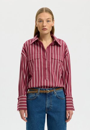 Žena s rovnými blond vlasmi má na sebe bordovú košeľu s bielymi zvislými pruhmi, modré džínsy a hnedý opasok, stojí pred jednofarebným pozadím.