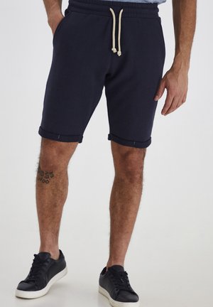 Shorts en coton bleu marine avec taille élastique, cordon de serrage ajustable et ourlets retroussés. Paire de baskets noires avec semelles blanches.