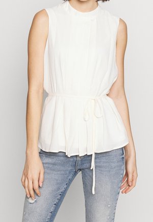 Kobieta w białej bluzce bez rękawów z wiązaniem w talii i jasnoniebieskich rozdartych dżinsach, stojąca na jednolitym tle.