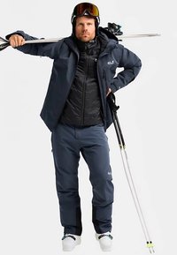 Skibekleidung in Marineblau, bestehend aus einer leichten Steppjacke, wasserdichten Hosen und weißen Skischuhen. Enthält Skistöcke und Skibrille.