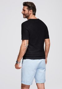 Camicetta nera a maniche corte con texture abbinata a pantaloni corti azzurri arrotolati. Tatuaggio visibile sul braccio, orologio argento al polso. Design semplice e casual.