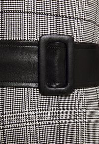 Ceinture en cuir noir avec une boucle rectangulaire, associée à un tissu à motif pied-de-poule noir et blanc, mettant en valeur des lignes nettes et une texture.