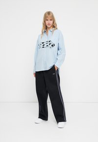 Light blue striped oversized shirt with black and white graphic design. Worn with loose black pants featuring a white stripe, and white shoes.