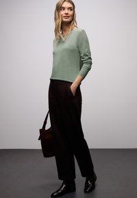 Zielony długi top z dekoltem w serek, zestawiony z czarnymi szerokimi spodniami. Modelka trzyma brązową skórzaną torbę na ramię i nosi czarne botki.