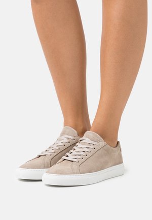 Baskets basses en daim beige avec semelles en caoutchouc blanc portées pieds nus, posées sur un fond gris clair uni.