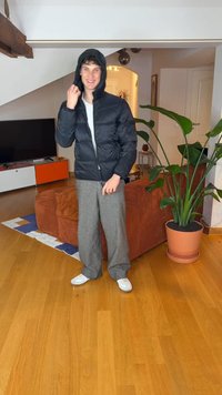 Svart puffjakke med hette, grå vide bukser og hvite joggesko. Bakgrunnen viser en potteplante og møbler.