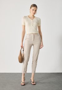 Haut à manches courtes côtelé crème avec un décolleté en V, associé à un pantalon beige ajusté. Le mannequin tient un sac à main beige et porte des sandales noires à lanières.