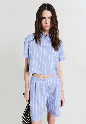 Camicia corta a maniche corte a righe blu chiaro con bottoni abbinata a pantaloni corti a vita alta con pieghe frontali e una texture liscia.
