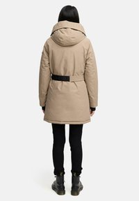 Parka beige avec une grande capuche et une ceinture noire. Tissu texturé et couleur unie, associé à un pantalon sombre et des bottes noires.