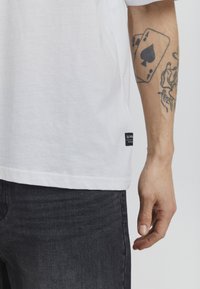 White cotton t-shirt with a square cut, short sleeves, and a black label at the hem. Paired with dark denim jeans and visible tattoos on the arm.