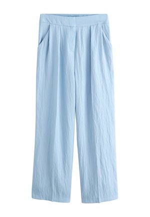 Donna in piedi contro una parete liscia che indossa un completo senza maniche azzurro chiaro con pantaloni a gamba larga e tacchi bianchi appuntiti, mano in tasca.