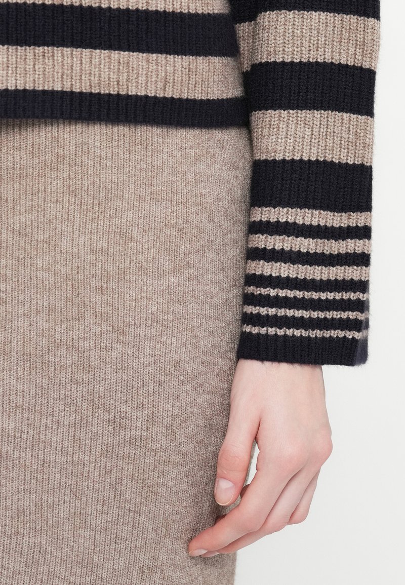 Pullover in Marineblau und Beige mit horizontalen Streifen, ausgestattet mit gerippten Bündchen. Beiger Rock aus einem weichen, strukturierten Stoff. Hand sichtbar.