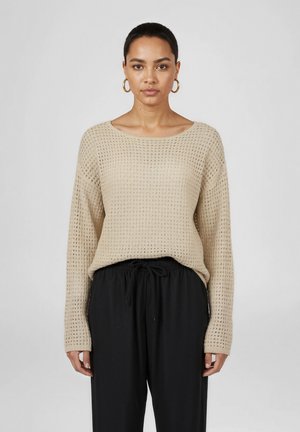 Kobieta stojąca przodem, ubrana w beżowy sweter o fakturze z długim rękawem, czarne spodnie ze ściągaczem oraz złote kolczyki koła na szarym tle.