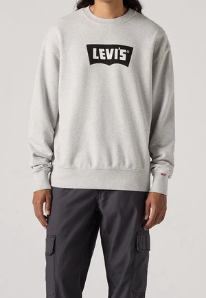 Osoba ubrana w jasnoszary sweter Levi's z czarnym logo i ciemnoszare spodnie typu cargo, stojąca na neutralnym tle.