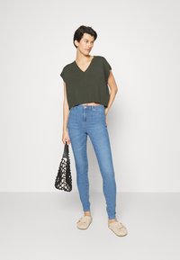 Olívazöld, V-kivágású, rövid fazonú top, kék skinny farmernadrág, bézs slip-on cipő és fekete horgolt táska. Egyszerű dizájn és laza illeszkedés.