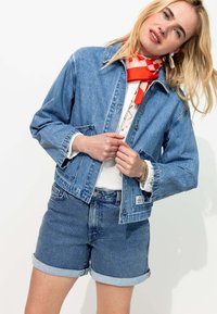 Veste en denim bleu clair avec un col, des poches avant et des boutons. Associée à un short en denim à ourlet roulé. Un foulard à carreaux rouges et blancs est porté.