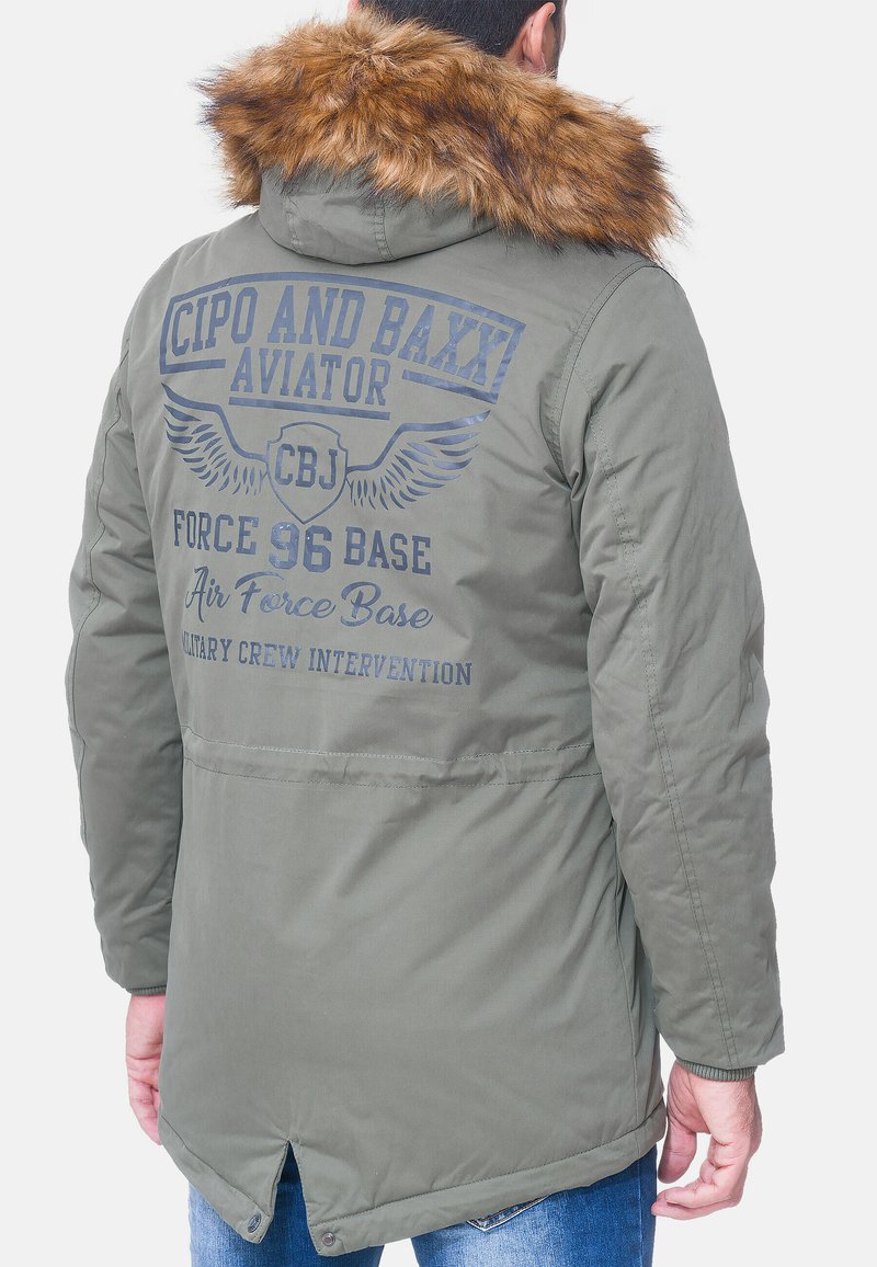 Giacca bomber verde con cappuccio foderato in pelliccia. Presenta testo stampato e grafiche di ali sul retro, con una finitura liscia e leggermente testurizzata.