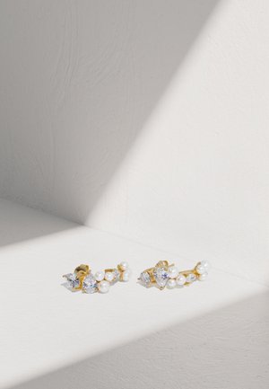 Orecchini d'oro con gemme trasparenti e perle bianche, dallo design delicato e forme miste. Sullo sfondo di una texture bianca.