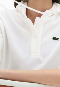 Vit pikétröja med texturerat tyg, med en krage med dragsko, en knappslå och en grön krokodillogotyp på vänster bröst.