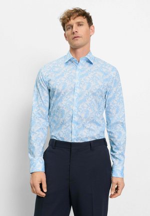 Mann, der ein hellblaues, langärmliges Hemd mit Knöpfen und einem blattartigen Muster sowie dunkelblaue Hosen trägt, steht vor einem schlichten weißen Hintergrund.