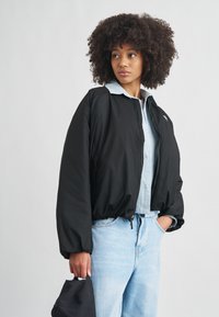 Czarna kurtka bomberka na suwak z elastycznymi mankietami, lekkiego materiału i luźnego kroju, zestawiona z jasnoniebieską koszulą z kołnierzykiem i dżinsami.