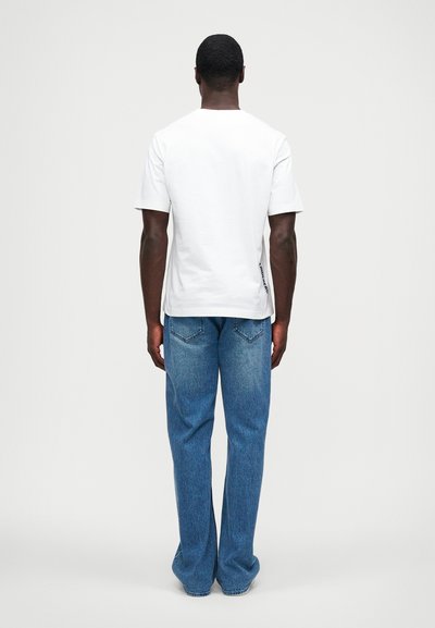 Balta medvilninė T marškinė su trumpomis rankovėmis, matoma iš nugaros; jie derinami su mėlynomis džinsinėmis kelnėmis, turinčiomis tiesų kirpimą ir kontrastingą siuvinėjimą.