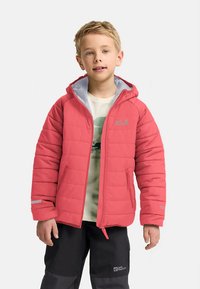 Jaunas berniukas, vilkintis raudoną kapučiną pūkinę striukę virš baltos marškinėlių su piešiniu ir juodų kelnių priešais vientisą baltą foną.