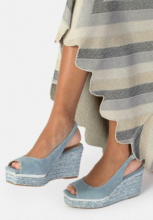 Piernas con sandalias cuña de ante azul claro con puntera abierta y tacones trenzados, parcialmente cubiertas por una falda a rayas con volantes.