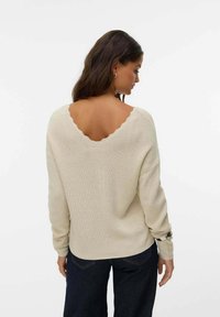 Maglione beige lavorato a maglia con scollatura a V ondulata, maniche lunghe e trama a costine. Indossato con jeans scuri, visto da dietro.