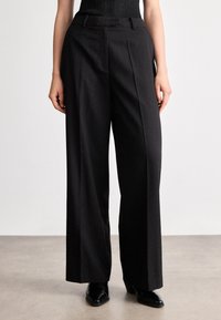 Melni pielāgoti bikses ar augstu jostu, krokām un brīvu piegriezumu. Izgatavotas no teksturēta materiāla ar smalkiem plankumiem un sānu kabatām.