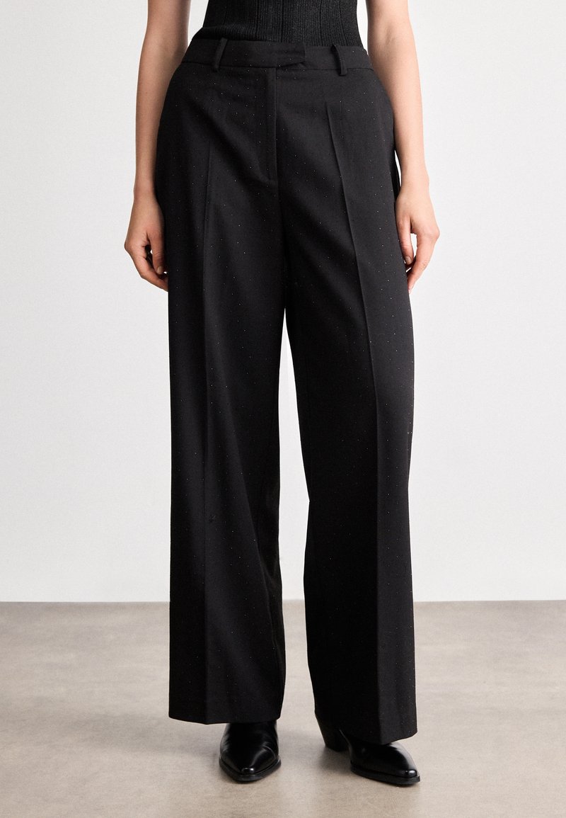 Melni pielāgoti bikses ar augstu jostu, krokām un brīvu piegriezumu. Izgatavotas no teksturēta materiāla ar smalkiem plankumiem un sānu kabatām.