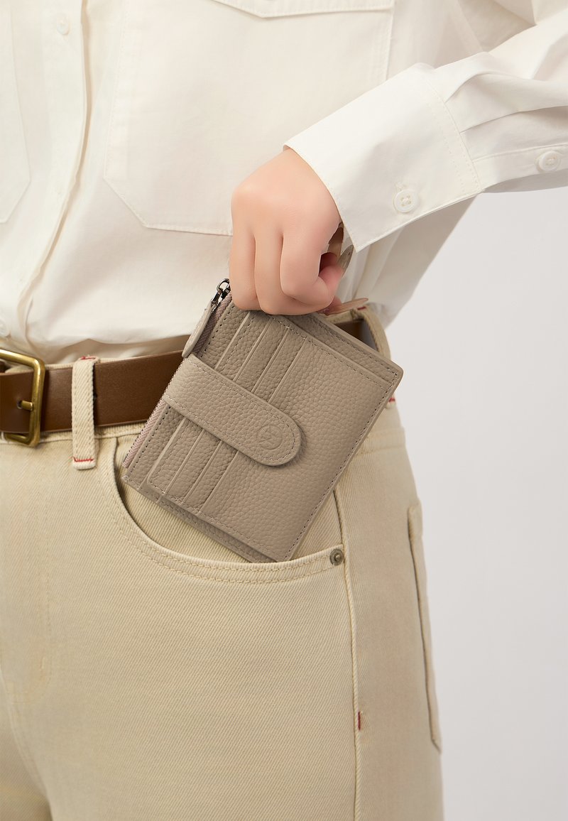 Beige leren portemonnee met kaartsleuven en een ritssluiting, gedeeltelijk gestopt in een lichtgekleurde denim zak. Close-up van portemonnee en zak.