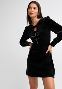 Schwarzes Samtkleid mit langen Ärmeln, gerafften Schultern, Schleifendetail am Ausschnitt und taillierter Silhouette. Glatte Textur und knielang.