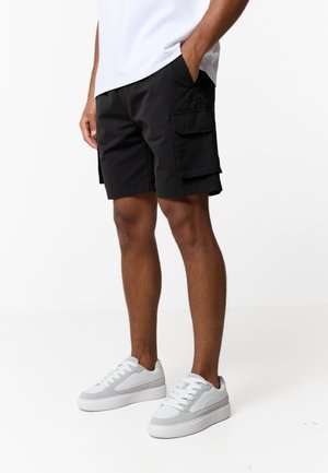 Mężczyzna w czarnych szortach cargo z klapkami na kieszeniach, białych tenisówkach i białej koszulce, stojący z rękami w kieszeniach na białym tle.