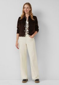Bruine corduroy jas met twee borstzakken, gedragen over een bloemenprint shirt. Crème kleurige wijde broek en zwarte platform schoenen maken de outfit compleet.