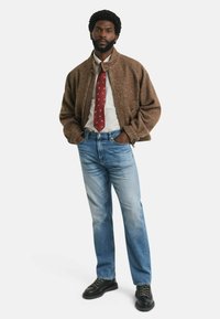 Bruine herringbone bomberjack, wit geruit overhemd, rode stropdas met kleine patronen, blauwe denim jeans en zwarte veterschoenen.