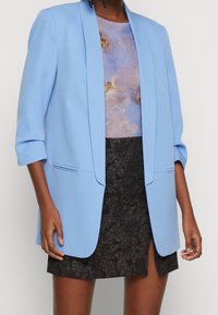Person wearing a light blue blazer, a blouse with cherub cloud print, and a black textured skirt with a front slit against a plain background.